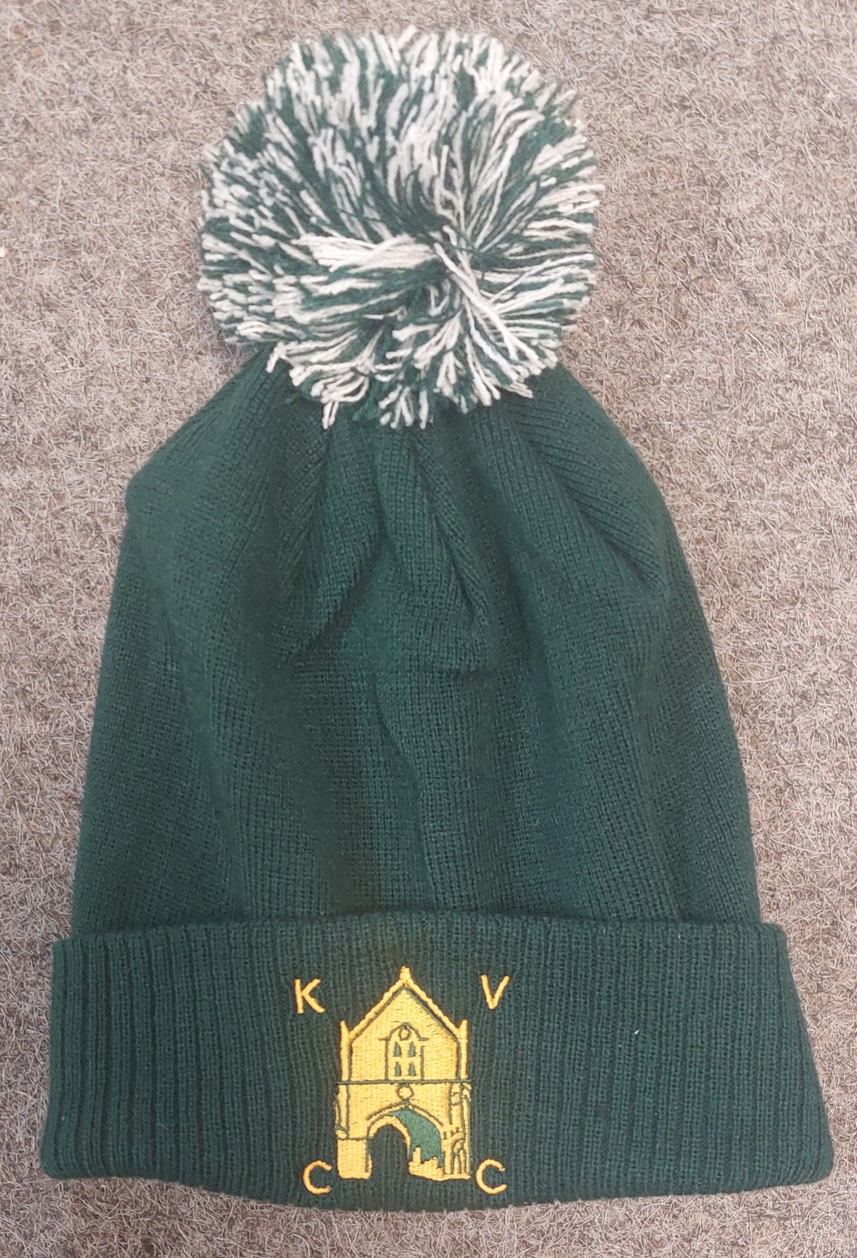 The Kingswood Village CC Snowstar Beanie (Bottle/Off White) by Martin Berrill Sports features dark green knit, a white and green pom-pom, an embroidered yellow building, and the Kingswood Village Club Logo on the front cuff. Laid flat on gray fabric.