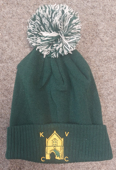 The Kingswood Village CC Snowstar Beanie (Bottle/Off White) by Martin Berrill Sports features dark green knit, a white and green pom-pom, an embroidered yellow building, and the Kingswood Village Club Logo on the front cuff. Laid flat on gray fabric.