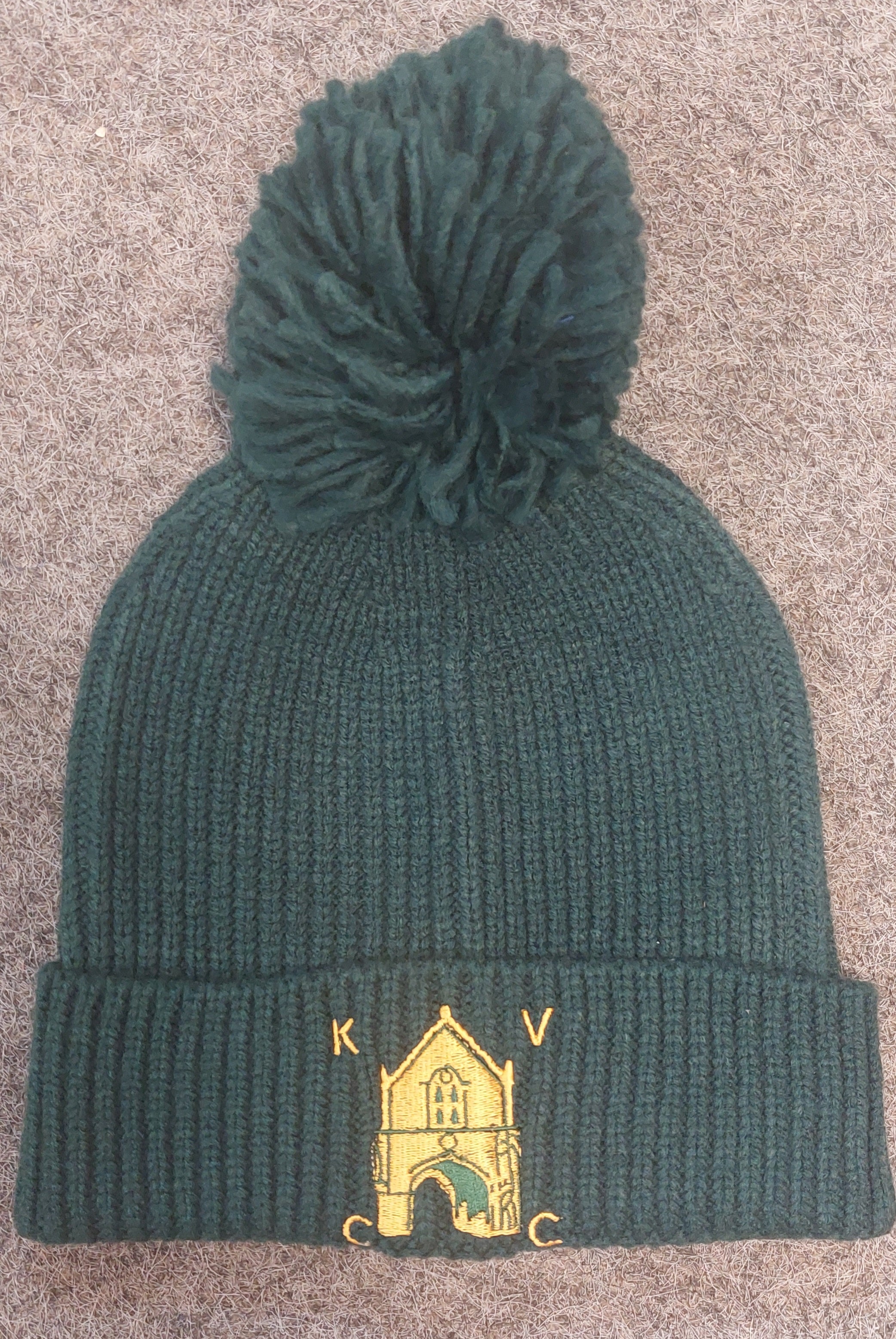 The Kingswood Village CC Knit Ribbed Beanie by Martin Berrill Sports is a dark green, pom-pom beanie with yellow embroidery of a building and "KVC" on the folded brim, shown on a textured gray surface.