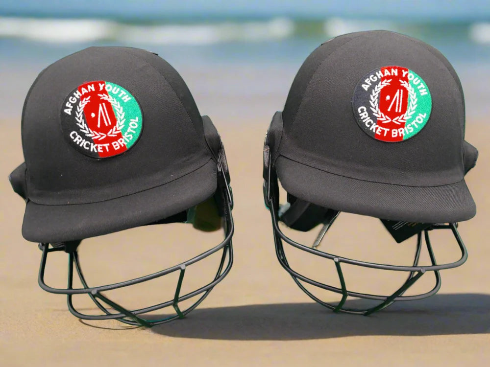 Two Custom Shrey Cricket Helmets with Club Logo by Shrey rest side by side on sand, with a blurred blue ocean and sky in the background.