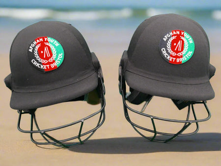 Two Custom Shrey Cricket Helmets with Club Logo by Shrey rest side by side on sand, with a blurred blue ocean and sky in the background.