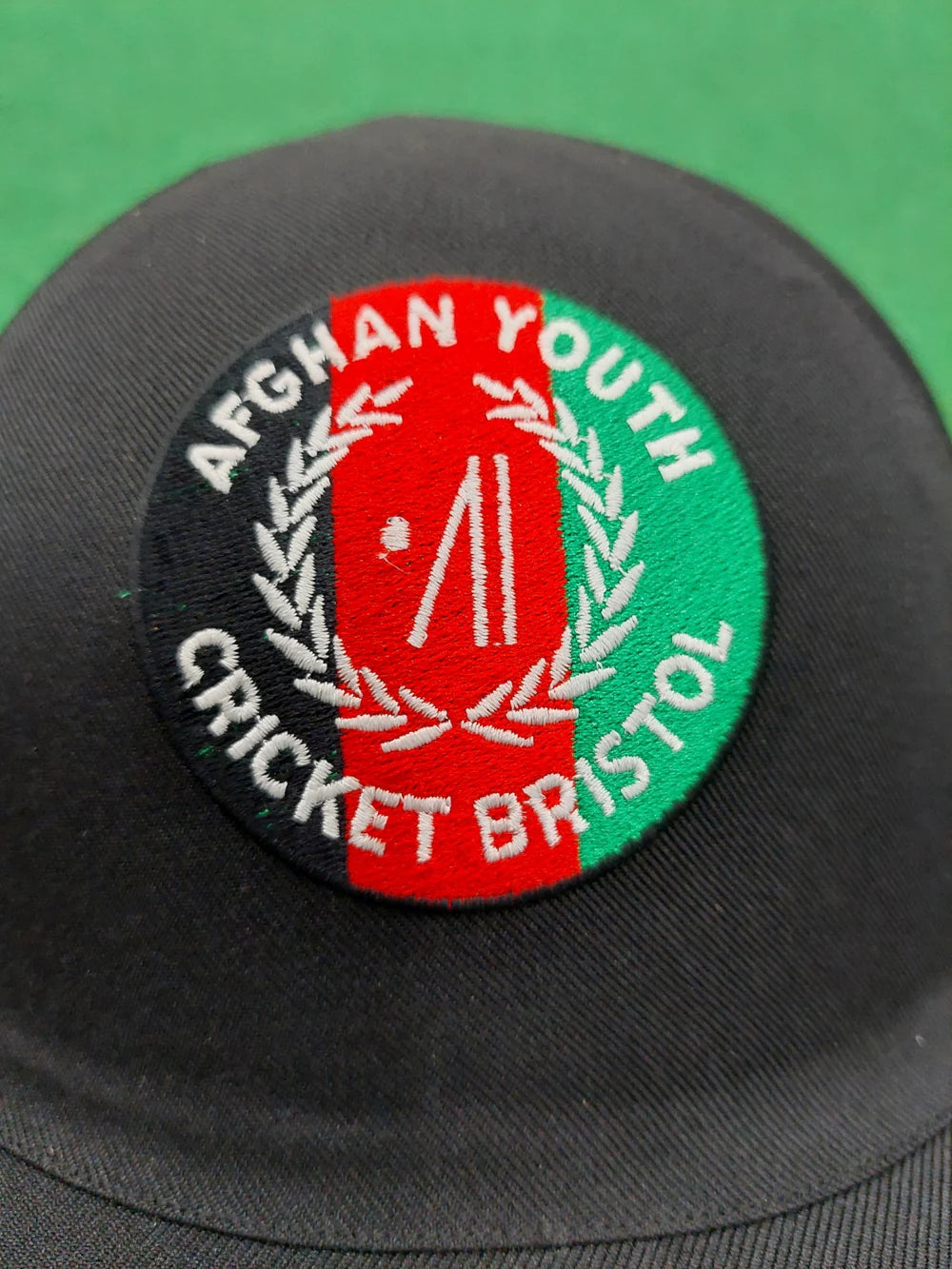 A black cap with a circular Club Logo patch—featuring "Afghan Youth Cricket Bristol," red and green background, white laurel, and cricket stumps—pairs perfectly with Custom Shrey Cricket Helmets by Shrey.