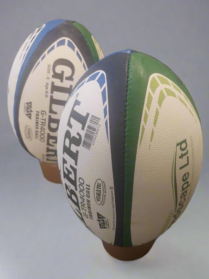 Two Custom Gilbert Match or Training Rugby Balls with Logos stand upright on brown tees; one is closer to the camera, both displaying Gilbert branding and custom panel designs in green, blue, and white.