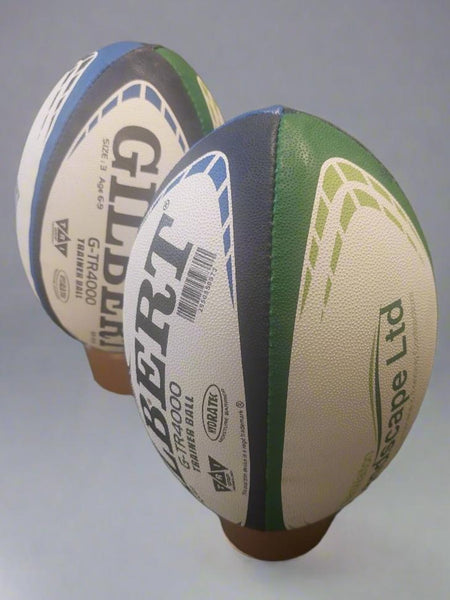 Two Custom Gilbert Match or Training Rugby Balls with Logos stand upright on brown tees; one is closer to the camera, both displaying Gilbert branding and custom panel designs in green, blue, and white.