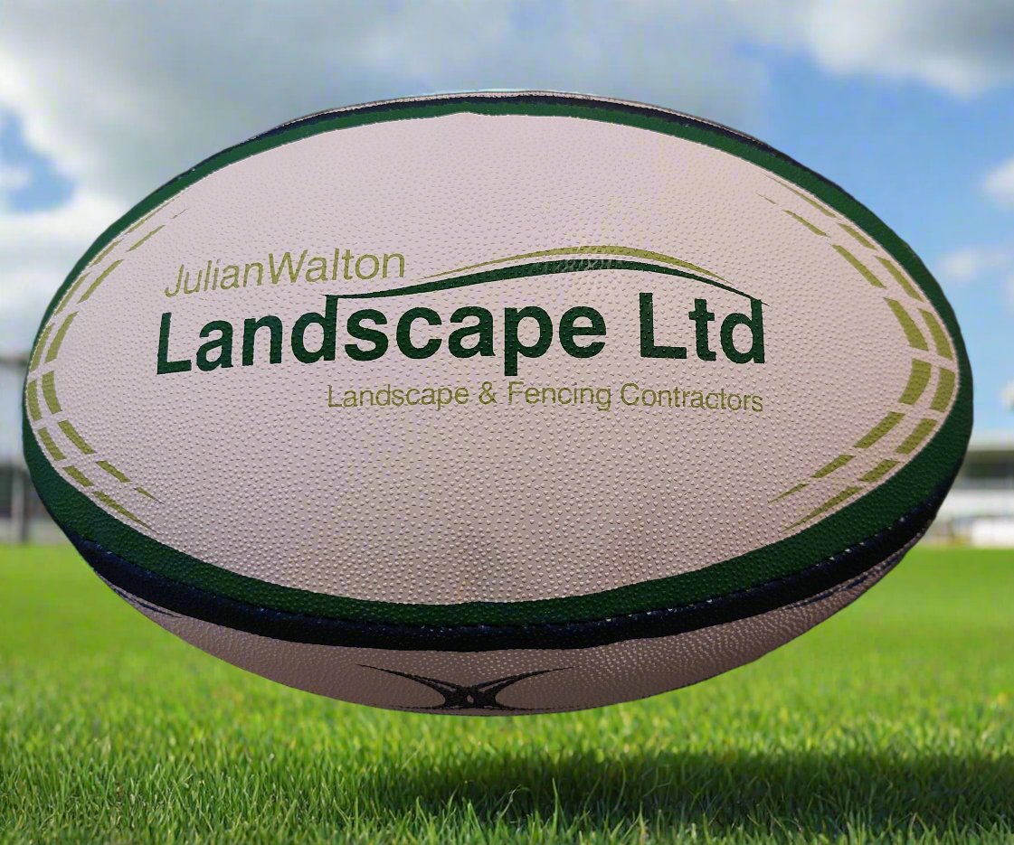 A Custom Gilbert Match or Training Rugby Ball with the Julian Walton Landscape Ltd logo and “Landscape & Fencing Contractors” printed on it rests on grass, with a blurred field and sky in the background.