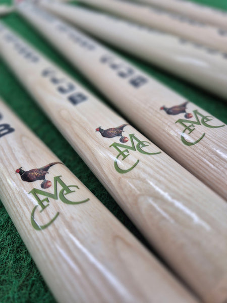 Several wooden cricket bats featuring a pheasant, cricket ball, and green MC logo are arranged on a green surface—perfectly paired with Gray Nicolls Custom Printed International Cricket Stumps for your next match.