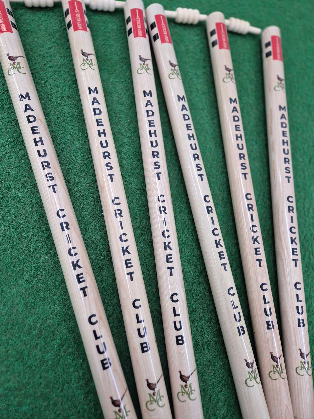 Six Gray Nicolls Custom Printed International Cricket Stumps on green turf, each with "MADEHURST CRICKET CLUB" in blue and a small deer on a bicycle image near the top.