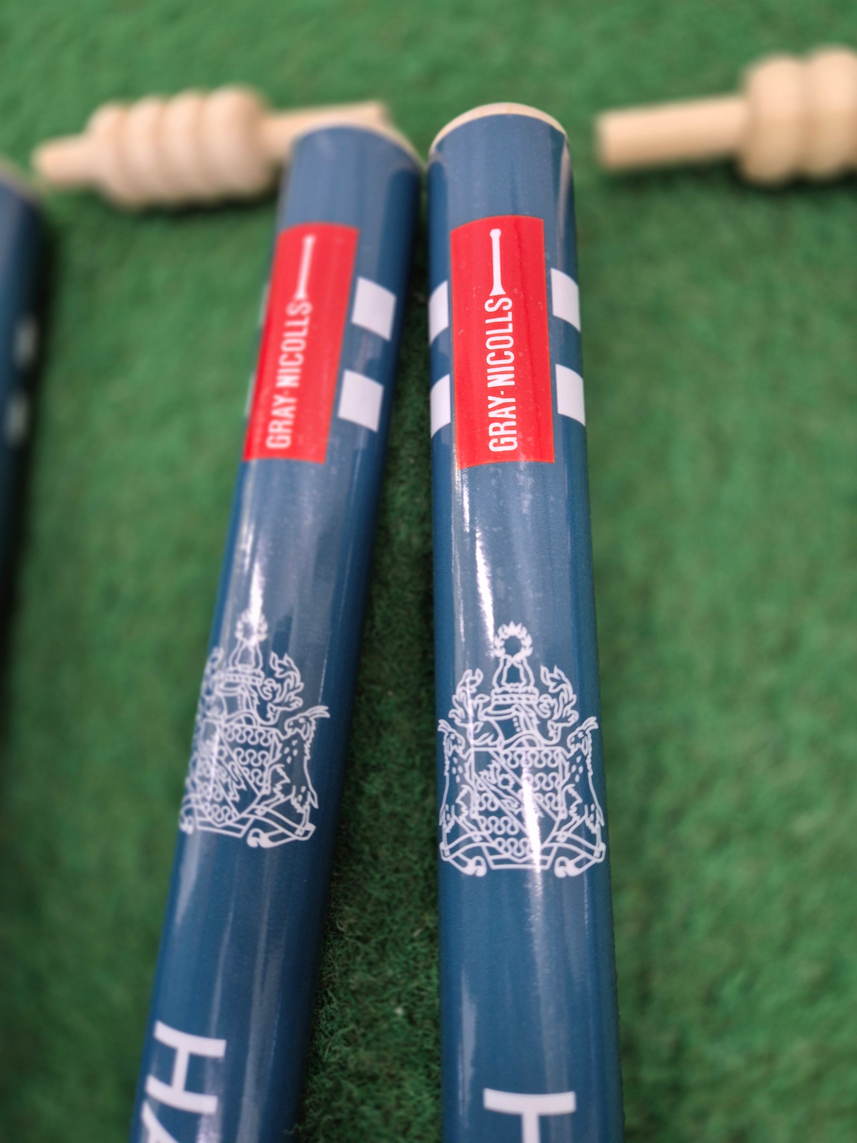 Two blue Custom Printed Club Cricket Stumps by Gray Nicolls with a white coat of arms are lying on green artificial turf, with wooden bails nearby.