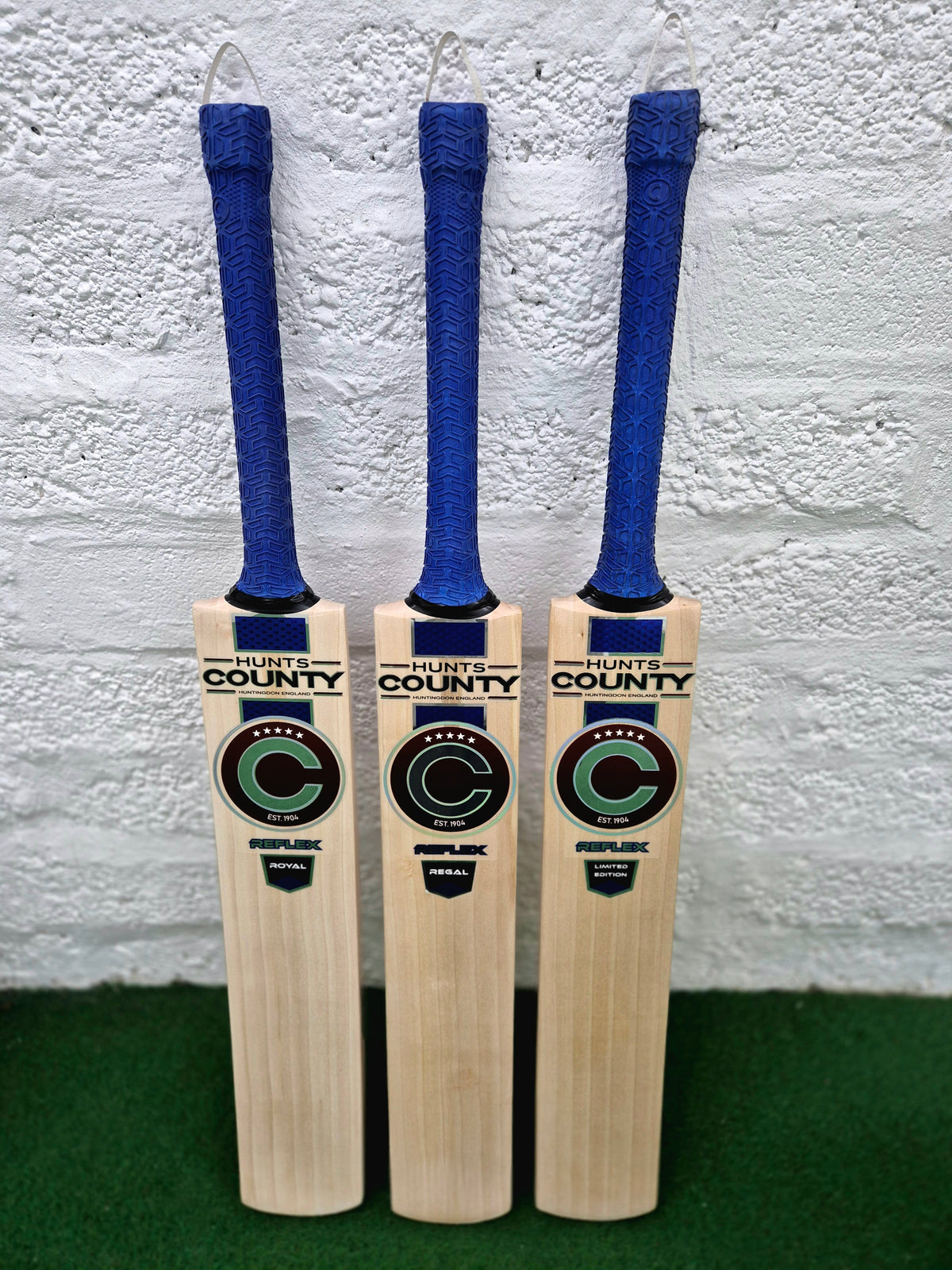 Three Hunts County Reflex Senior Cricket Bats, each with a blue grip, are displayed upright against a white wall. Models include the Reflex Royal, Reflex Regal, and Reflex Limited Edition by Hunts County.