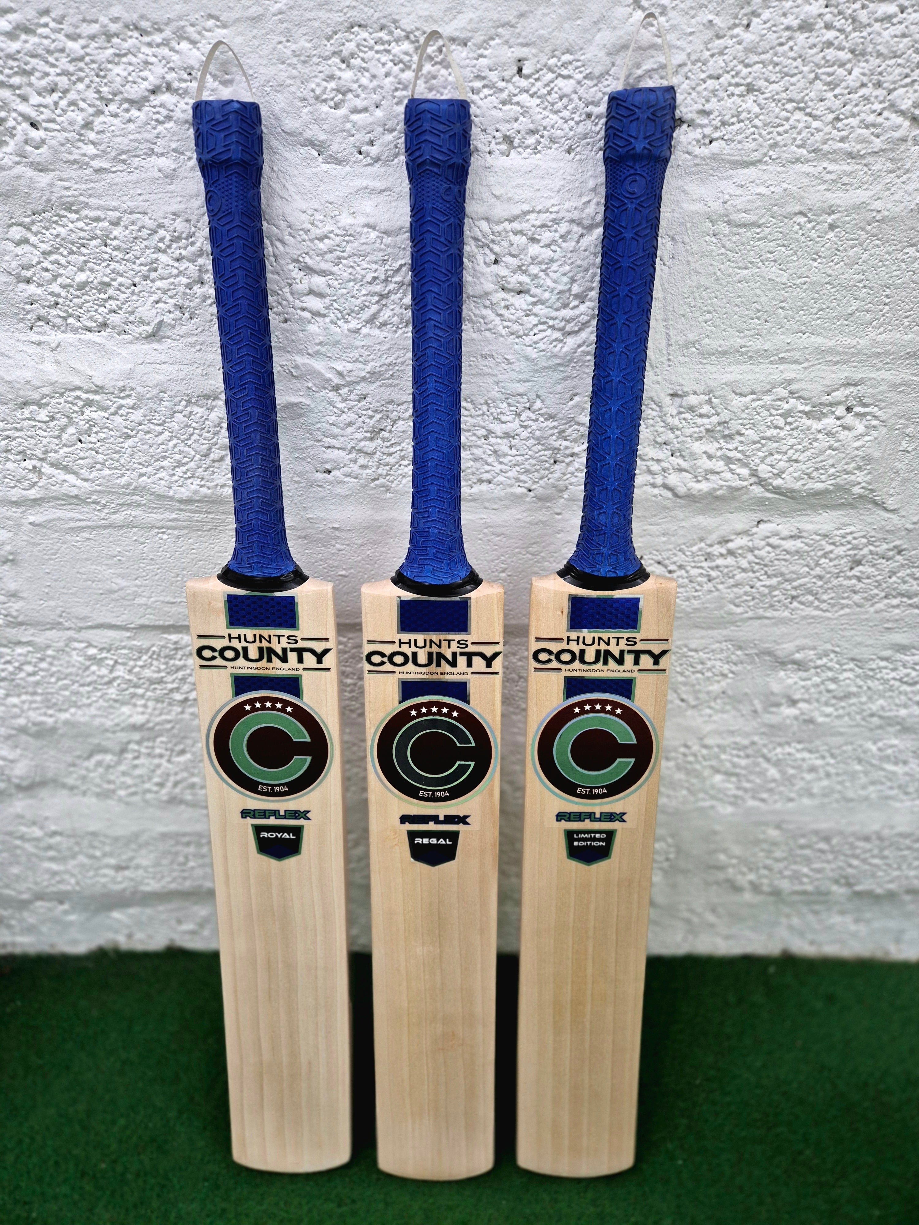 Three Hunts County Reflex Senior Cricket Bats, each with a blue grip, are displayed upright against a white wall. Models include the Reflex Royal, Reflex Regal, and Reflex Limited Edition by Hunts County.