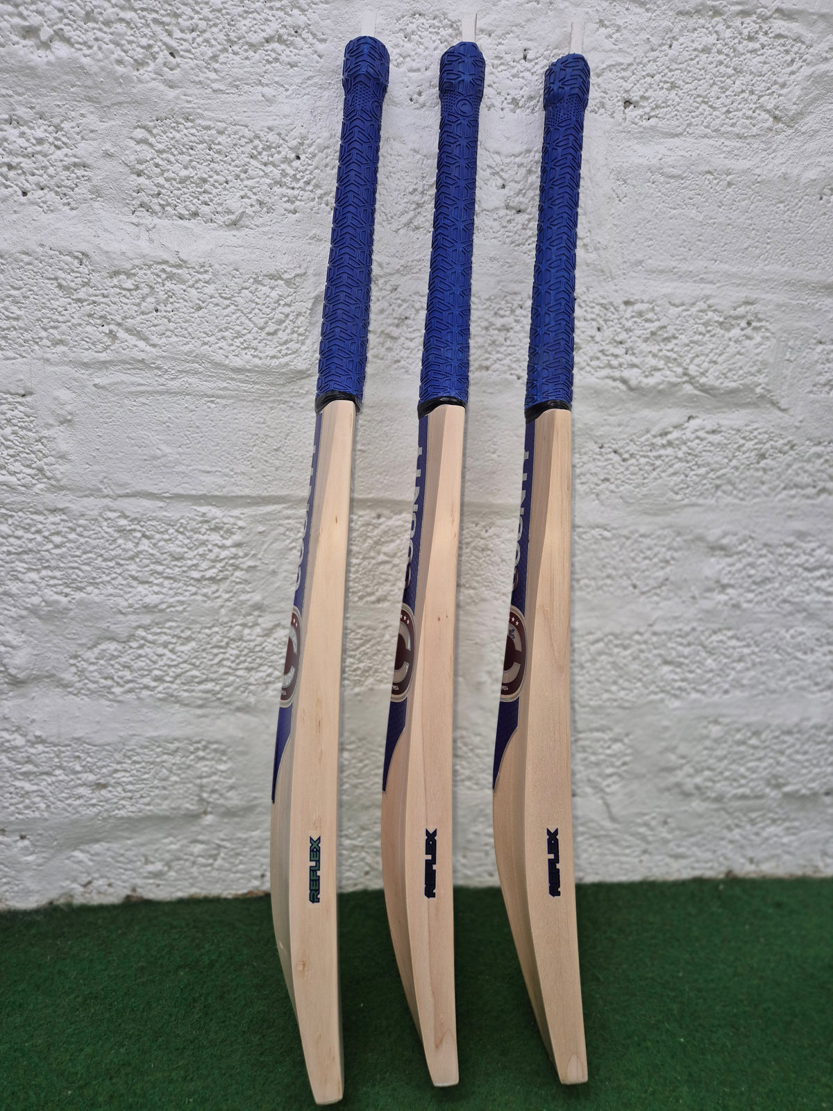 Three Hunts County Reflex Senior Cricket Bats with blue handles stand upright on green turf against a white brick wall—a top pick for senior players by Hunts County.