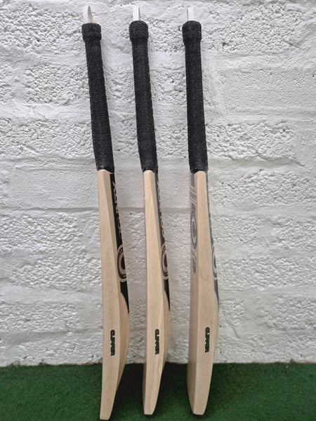 Three Hunts County Clipper Senior Cricket Bats by Hunts County, made from English willow and featuring black handles, are standing upright on a green carpet, leaning against a white brick wall.