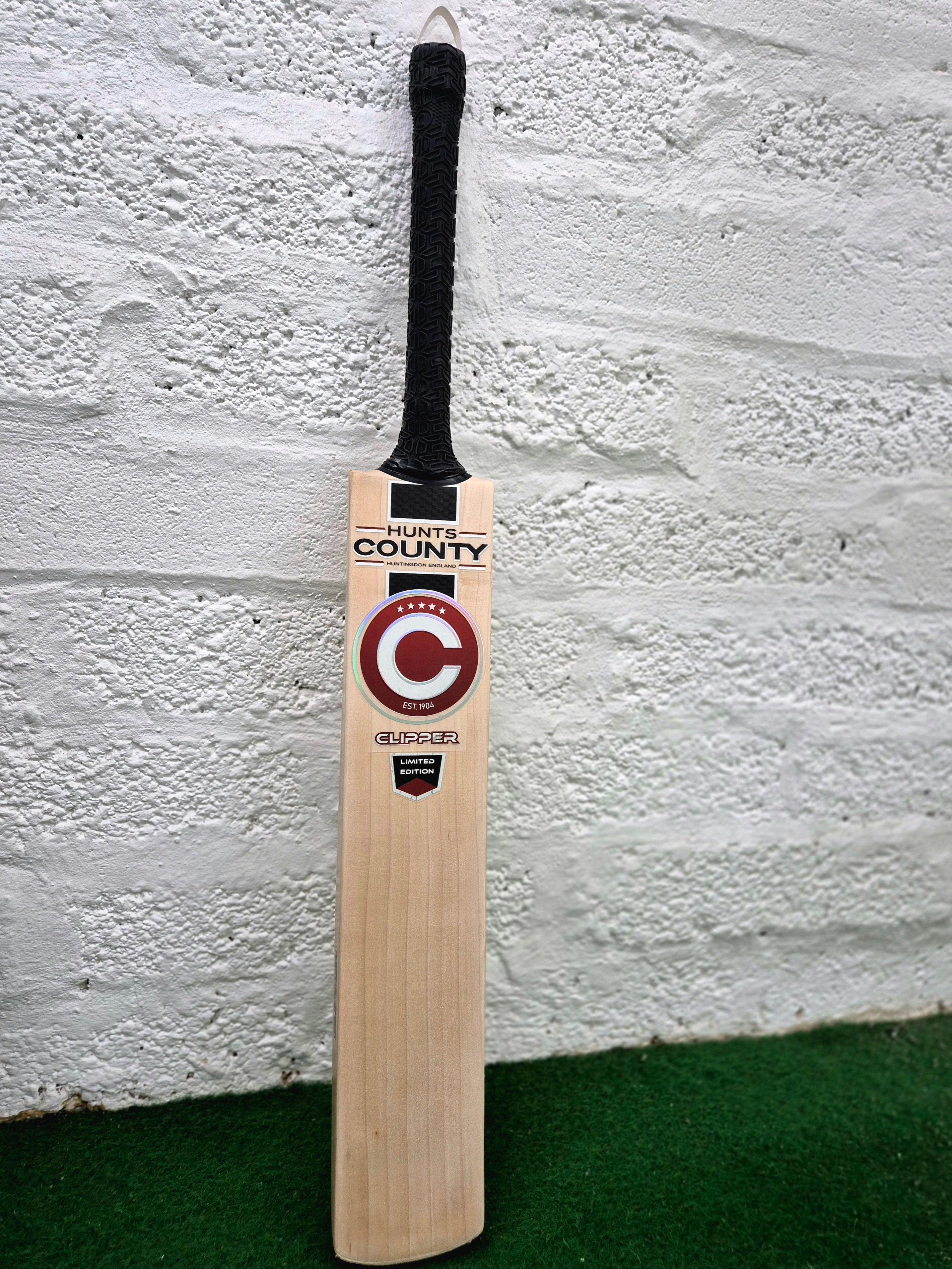 A Hunts County Clipper Senior Cricket Bat by Hunts County, featuring a black handle, stands upright against a white textured wall on green turf. Made from premium English willow, the bat showcases the signature Hunts County Clipper branding and a bold C logo on its front.