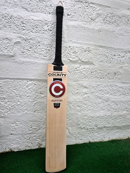 A Hunts County Clipper Senior Cricket Bat by Hunts County, featuring a black handle, stands upright against a white textured wall on green turf. Made from premium English willow, the bat showcases the signature Hunts County Clipper branding and a bold C logo on its front.