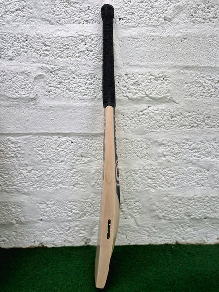 A Hunts County Clipper Senior Cricket Bat by Hunts County, made from premium English willow and featuring a black handle, is standing upright against a white textured brick wall on a green carpeted floor.