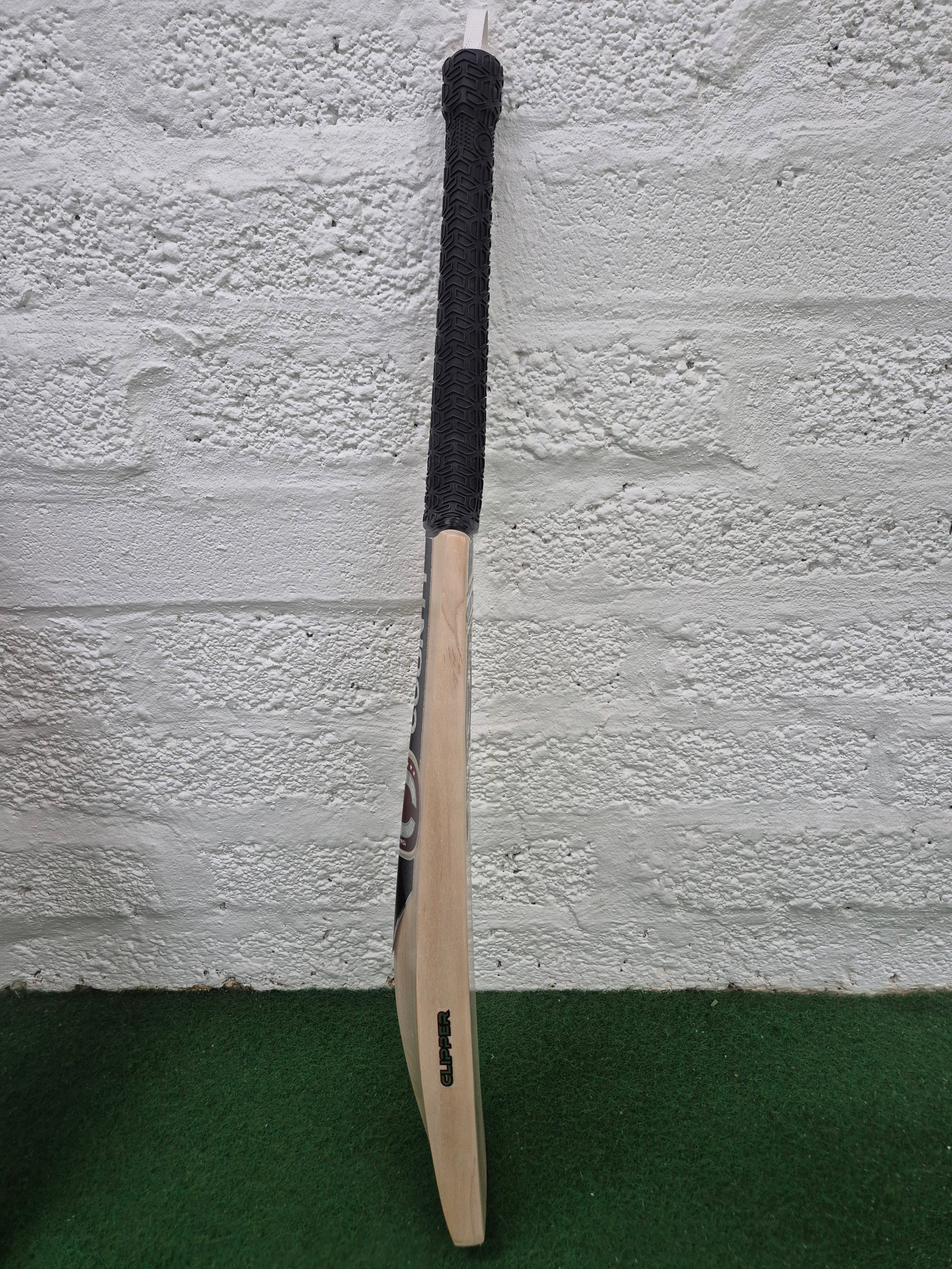 A Hunts County Clipper Senior Cricket Bat by Hunts County, crafted from quality English willow and equipped with a black handle grip, stands upright against a white textured brick wall on a green carpet.