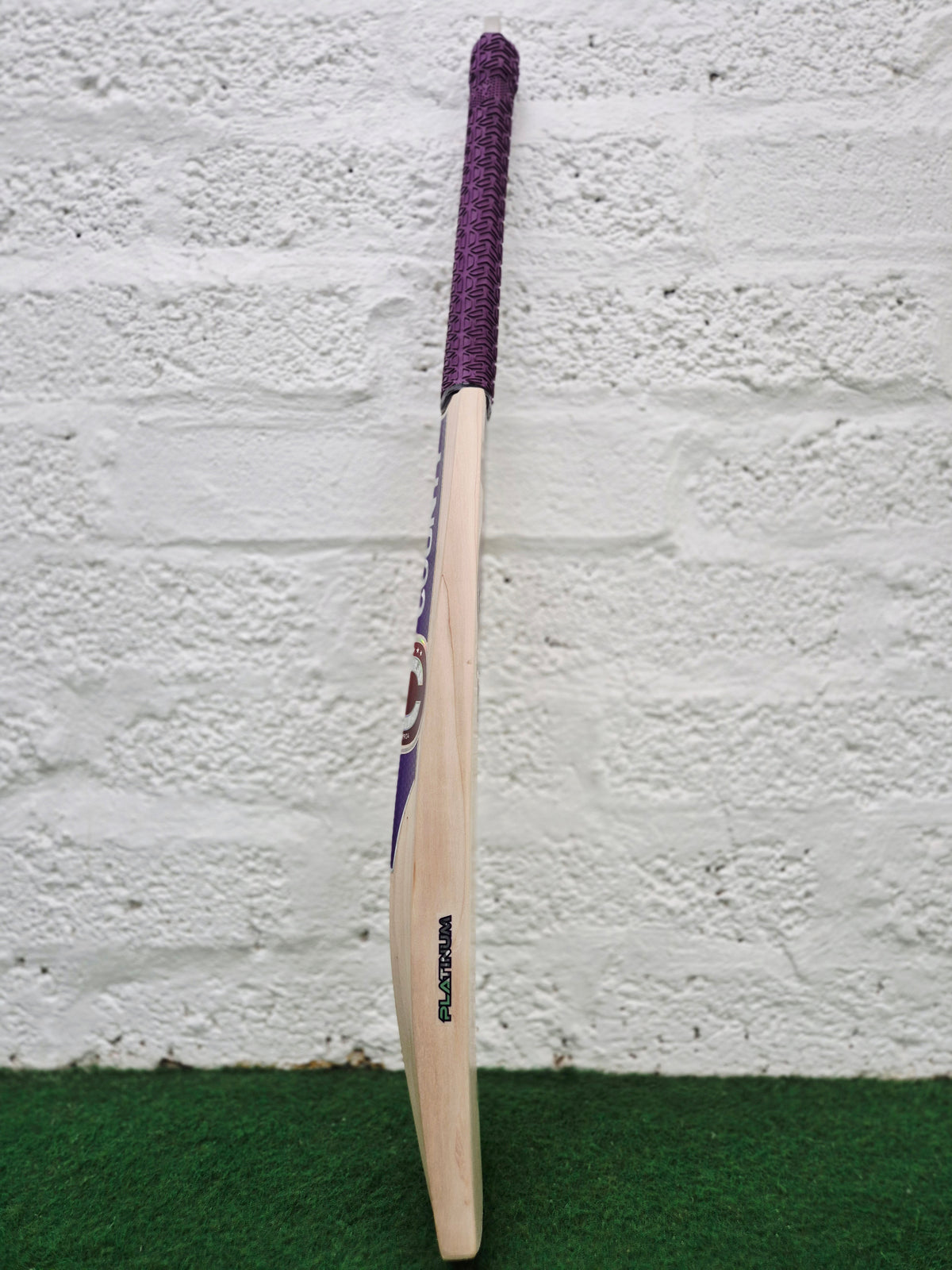 A Hunts County Platinum Players Senior Cricket Bat with a purple grip stands upright on green turf against a white brick wall.