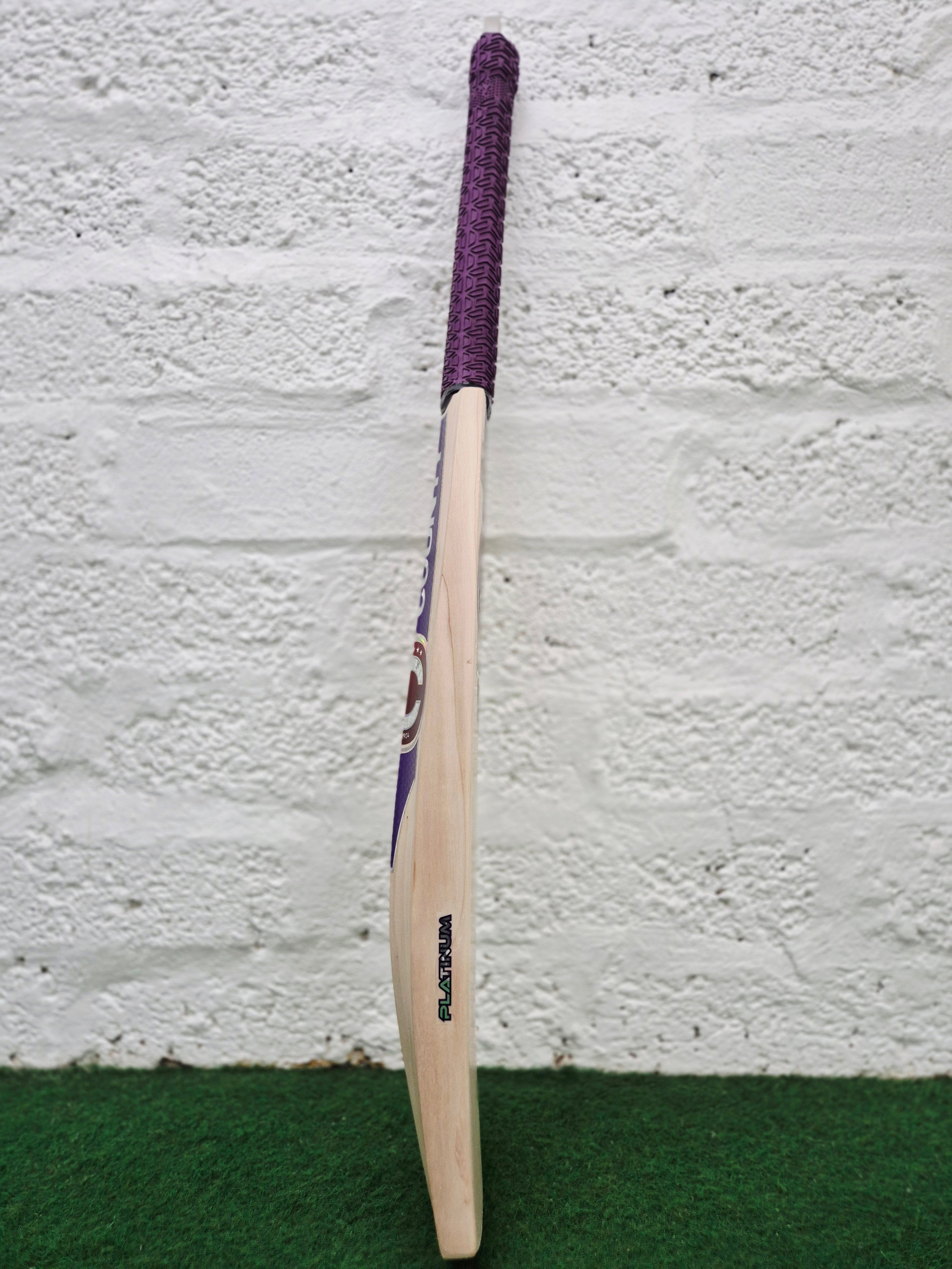A Hunts County Platinum Players Senior Cricket Bat with a purple grip stands upright on green turf against a white brick wall.