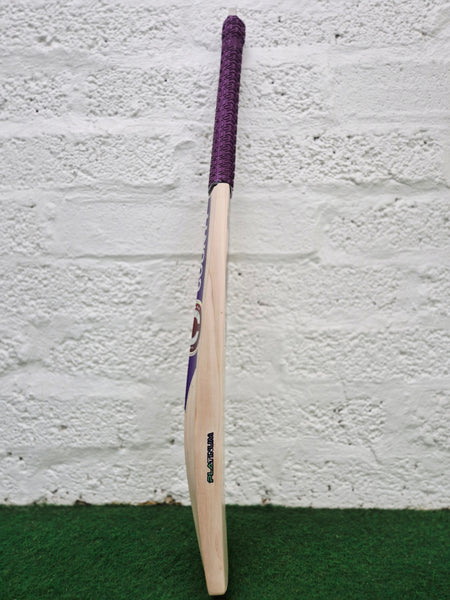 A Hunts County Platinum Players Senior Cricket Bat with a purple grip stands upright on green turf against a white brick wall.