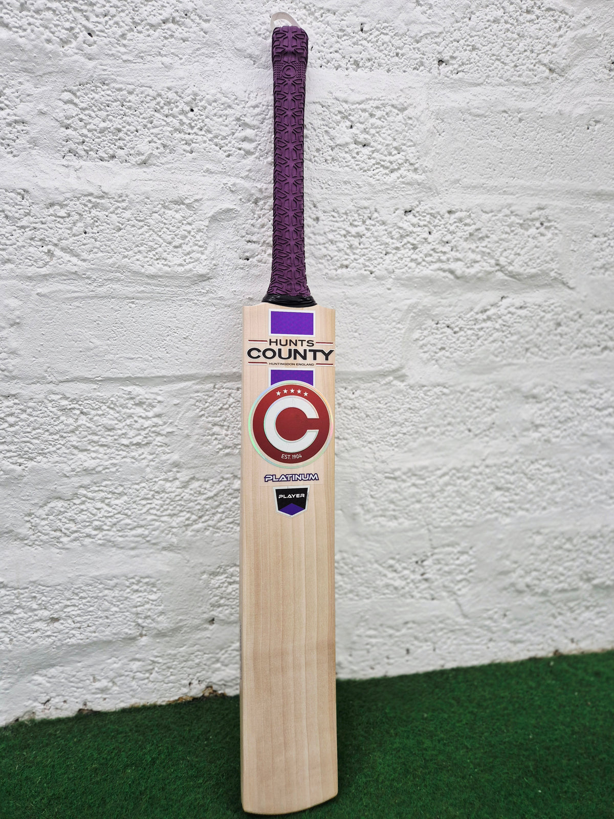 A Hunts County Platinum Players Senior Cricket Bat with a purple grip is standing upright against a white brick wall on a green surface.