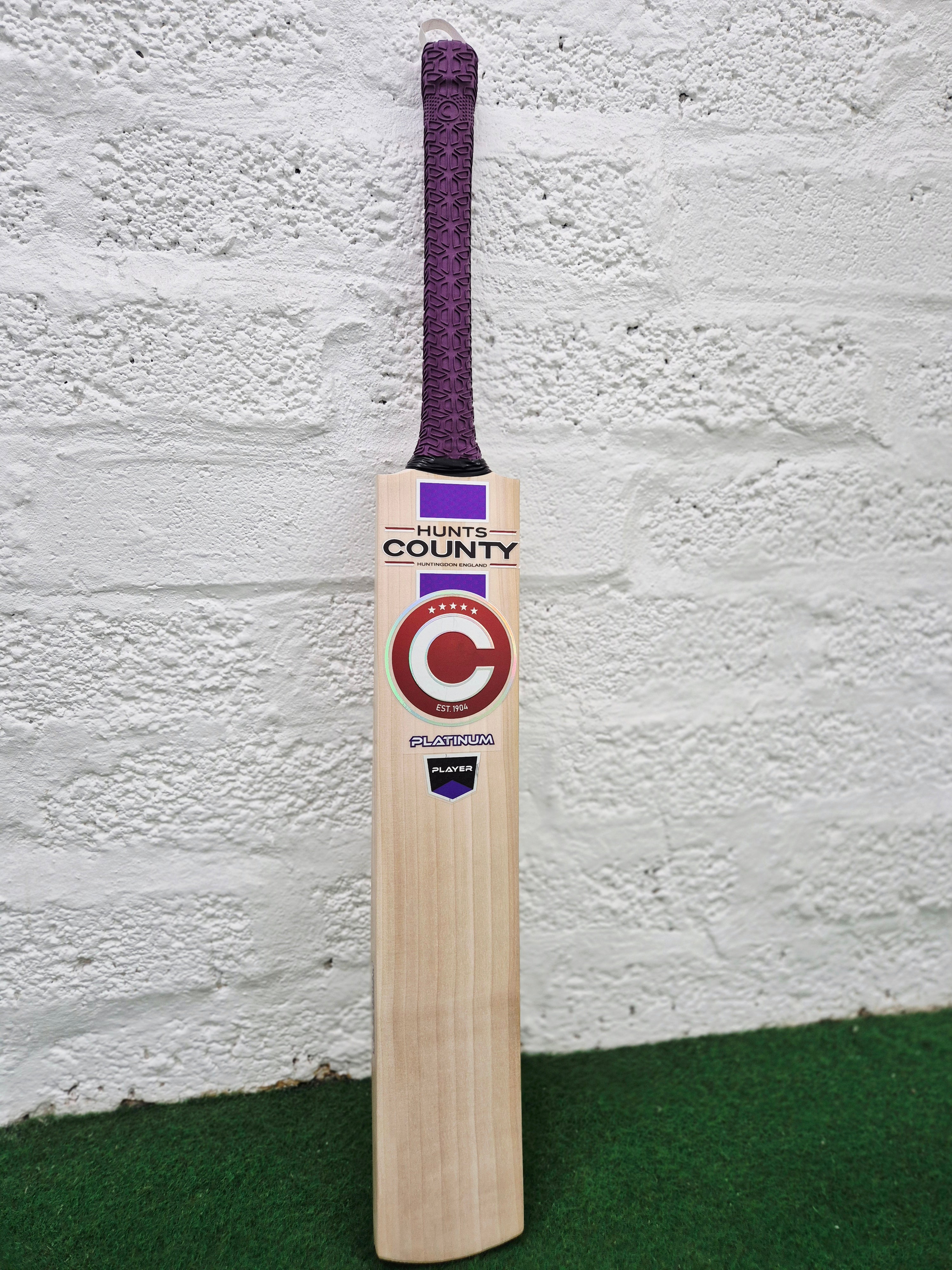 A Hunts County Platinum Players Senior Cricket Bat with a purple grip is standing upright against a white brick wall on a green surface.
