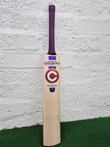 A Hunts County Platinum Players Senior Cricket Bat with a purple grip is standing upright against a white brick wall on a green surface.