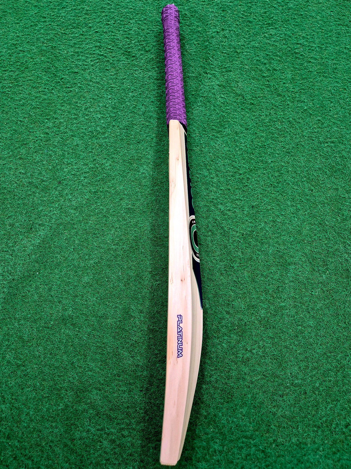 A Hunts County Platinum Players Senior Cricket Bat, featuring a purple grip, is positioned vertically with the handle at the top on a green surface.