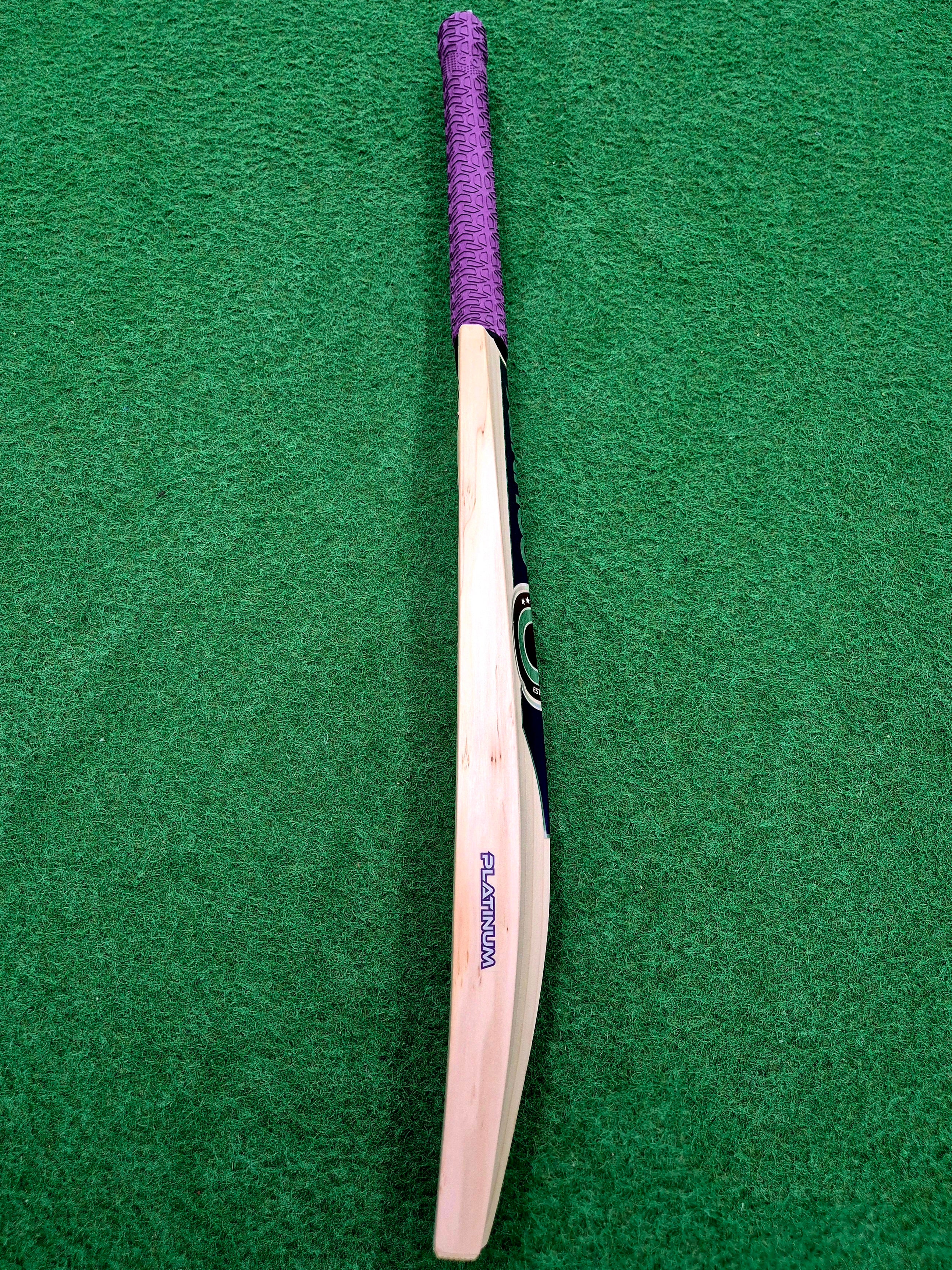A Hunts County Platinum Players Senior Cricket Bat, featuring a purple grip, is positioned vertically with the handle at the top on a green surface.