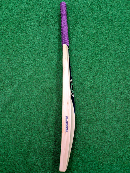 A Hunts County Platinum Players Senior Cricket Bat, featuring a purple grip, is positioned vertically with the handle at the top on a green surface.