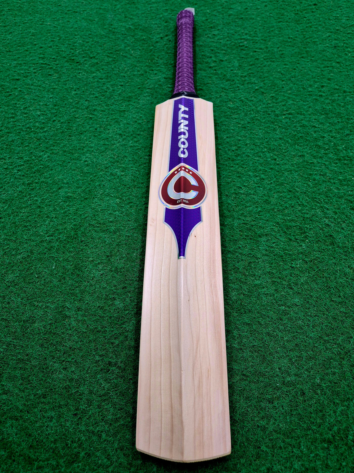 A Hunts County Platinum Players Senior Cricket Bat, crafted from English Willow and featuring a purple handle with COUNTY branding, lies on green artificial turf.