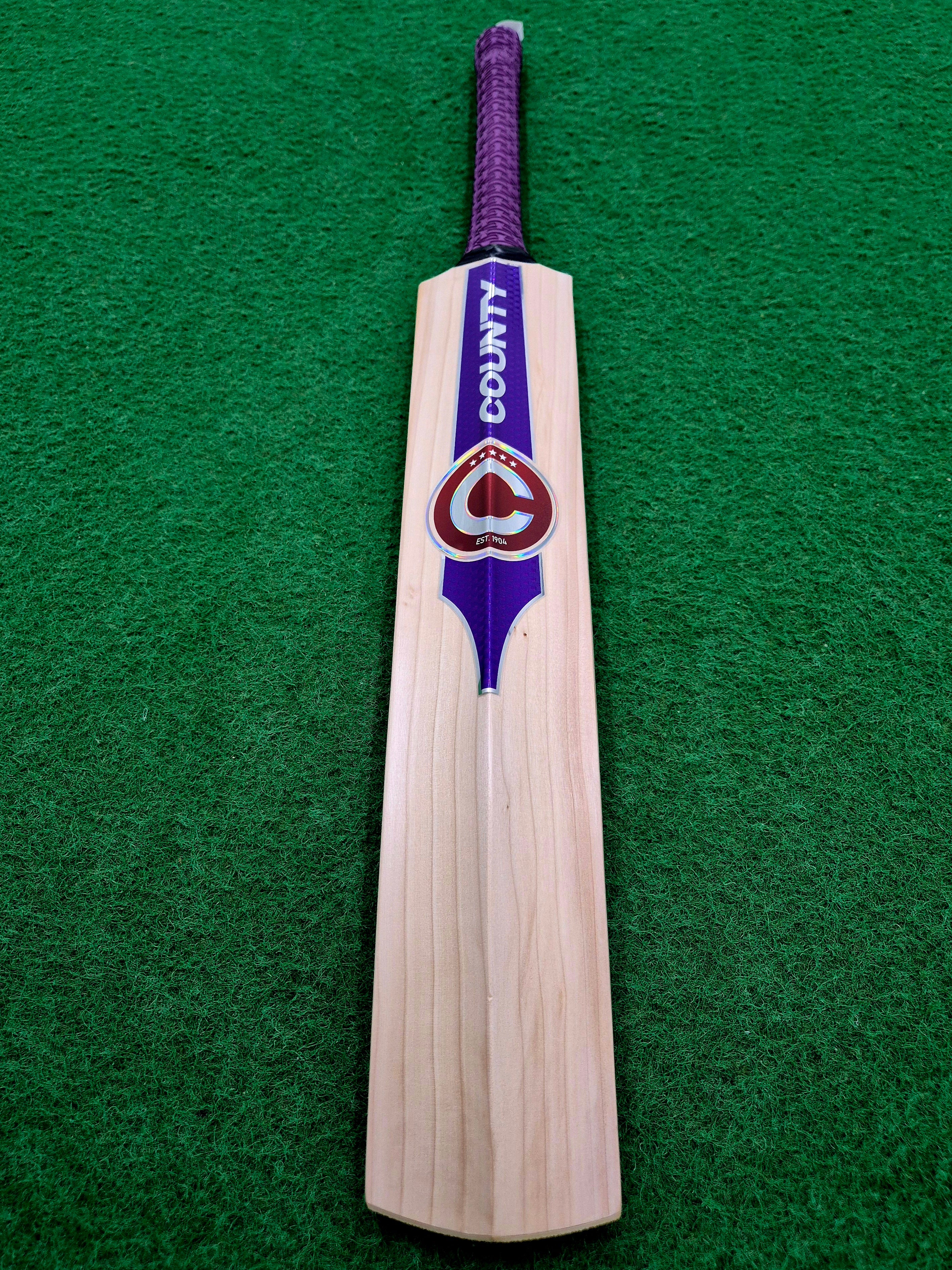 A Hunts County Platinum Players Senior Cricket Bat, crafted from English Willow and featuring a purple handle with COUNTY branding, lies on green artificial turf.