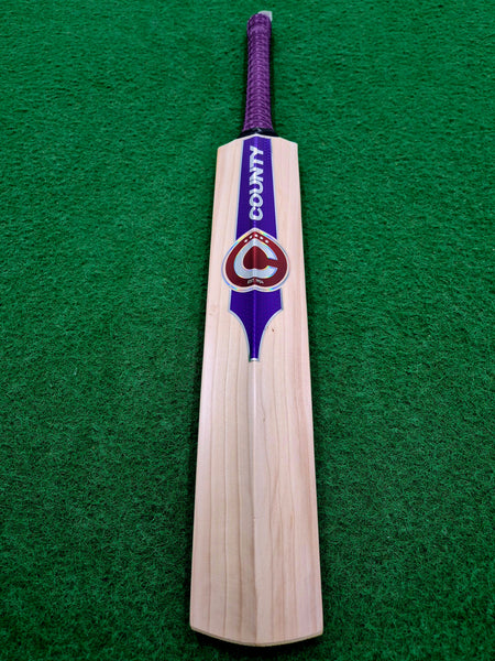 A Hunts County Platinum Players Senior Cricket Bat, crafted from English Willow and featuring a purple handle with COUNTY branding, lies on green artificial turf.