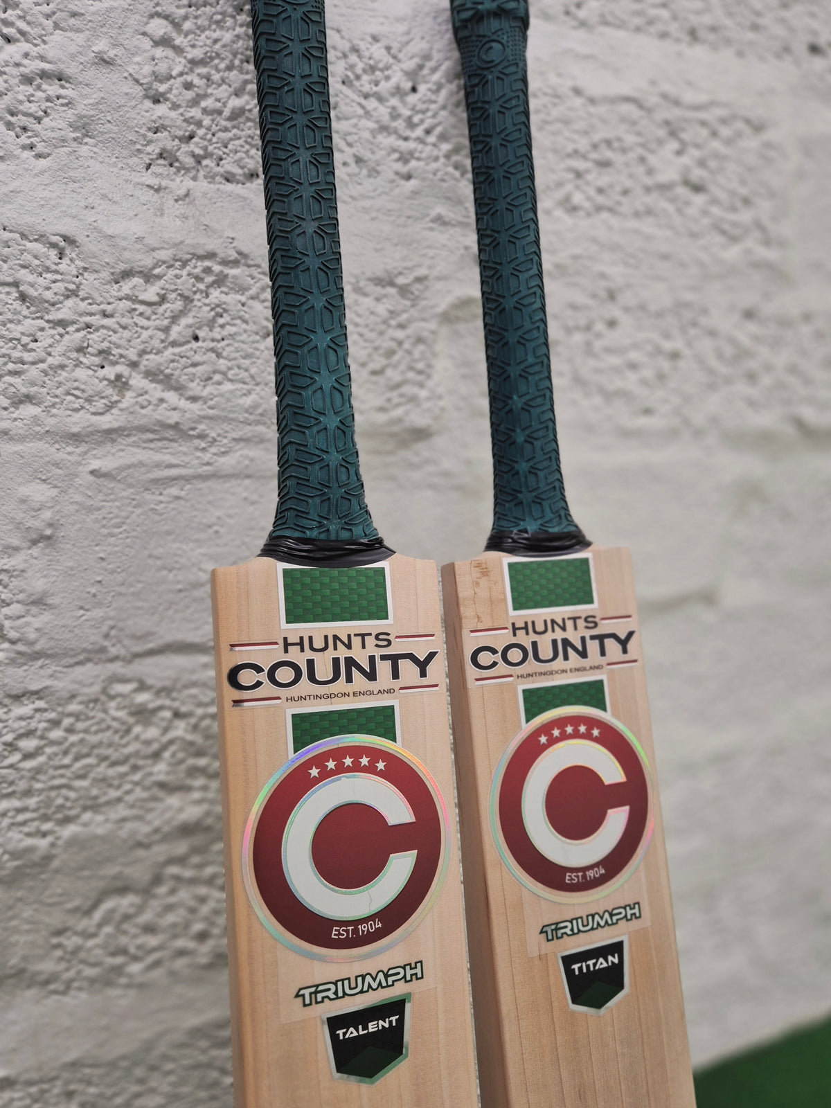 Two Hunts County Triumph Senior Cricket Bats with green grips rest against a white textured wall. Both bats feature a red and white circular Hunts County logo, with TRIUMPH prominently displayed at the bottom. Crafted from premium English Willow, each bat is engineered for serious players seeking superior performance in the 2026 senior cricket bat range.