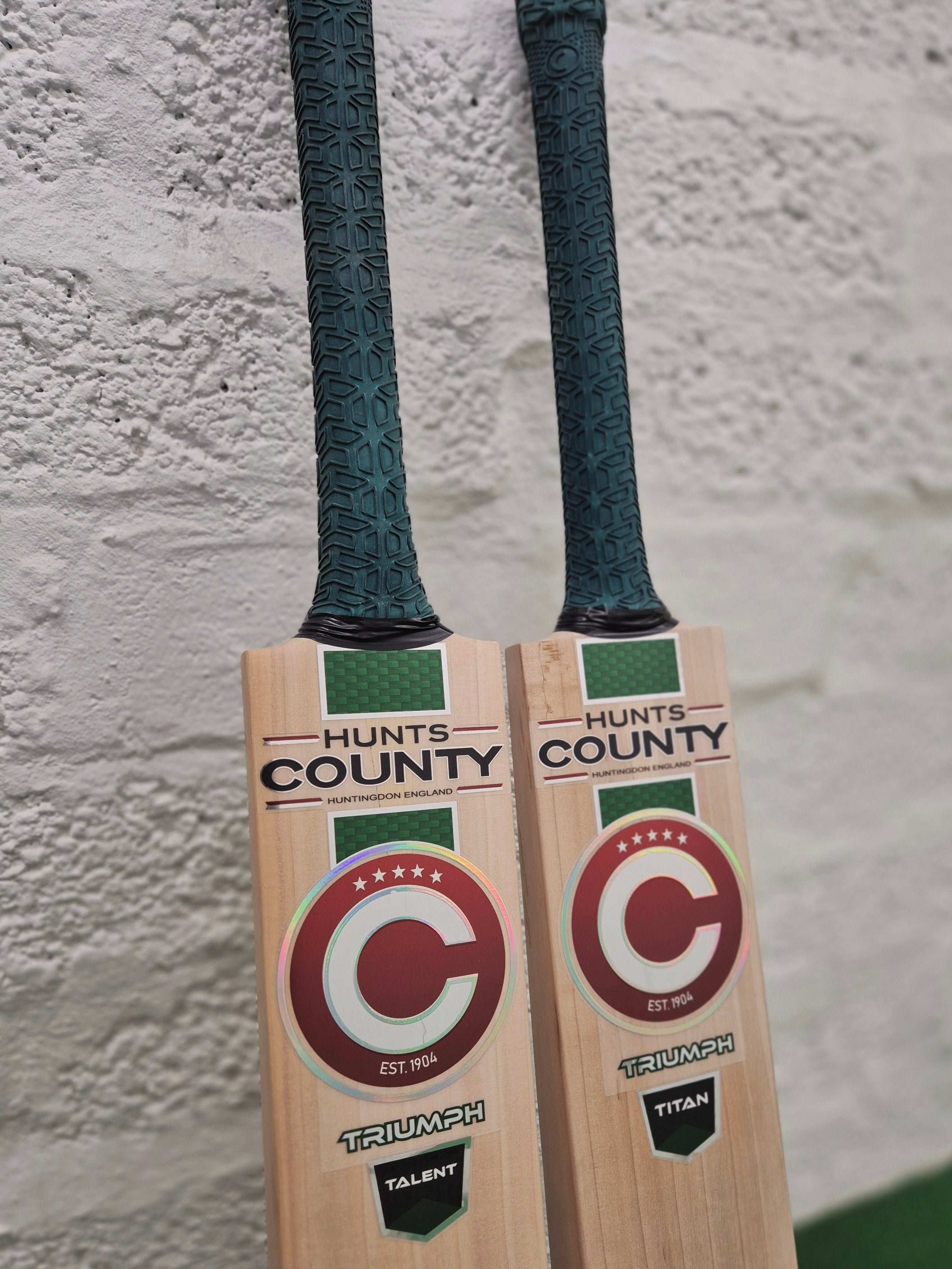 Two Hunts County Triumph Senior Cricket Bats with green grips rest against a white textured wall. Both bats feature a red and white circular Hunts County logo, with TRIUMPH prominently displayed at the bottom. Crafted from premium English Willow, each bat is engineered for serious players seeking superior performance in the 2026 senior cricket bat range.
