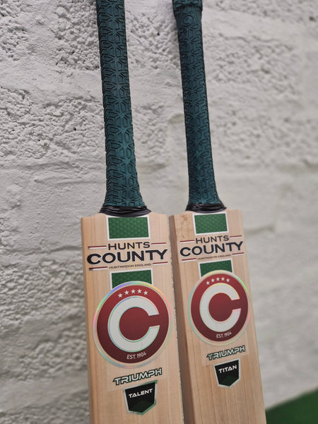 Two Hunts County Triumph Senior Cricket Bats with green grips rest against a white textured wall. Both bats feature a red and white circular Hunts County logo, with TRIUMPH prominently displayed at the bottom. Crafted from premium English Willow, each bat is engineered for serious players seeking superior performance in the 2026 senior cricket bat range.