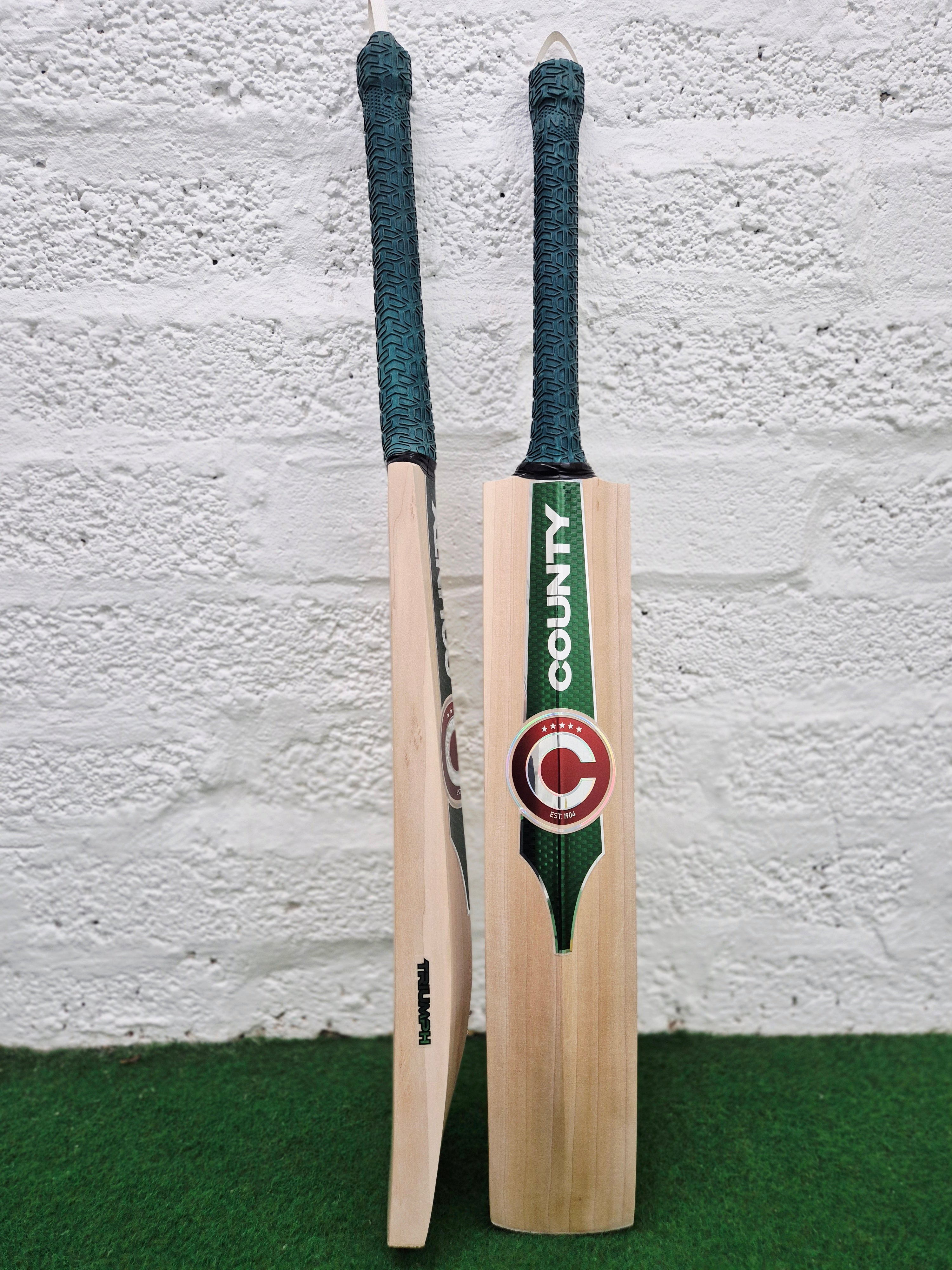 Two Hunts County Triumph Senior Cricket bats with green handles stand upright on artificial grass against a white brick wall. The front bat features the distinctive COUNTY logo, while the side view showcases the substantial thickness typical of a 2026 model senior English Willow bat from Hunts County.