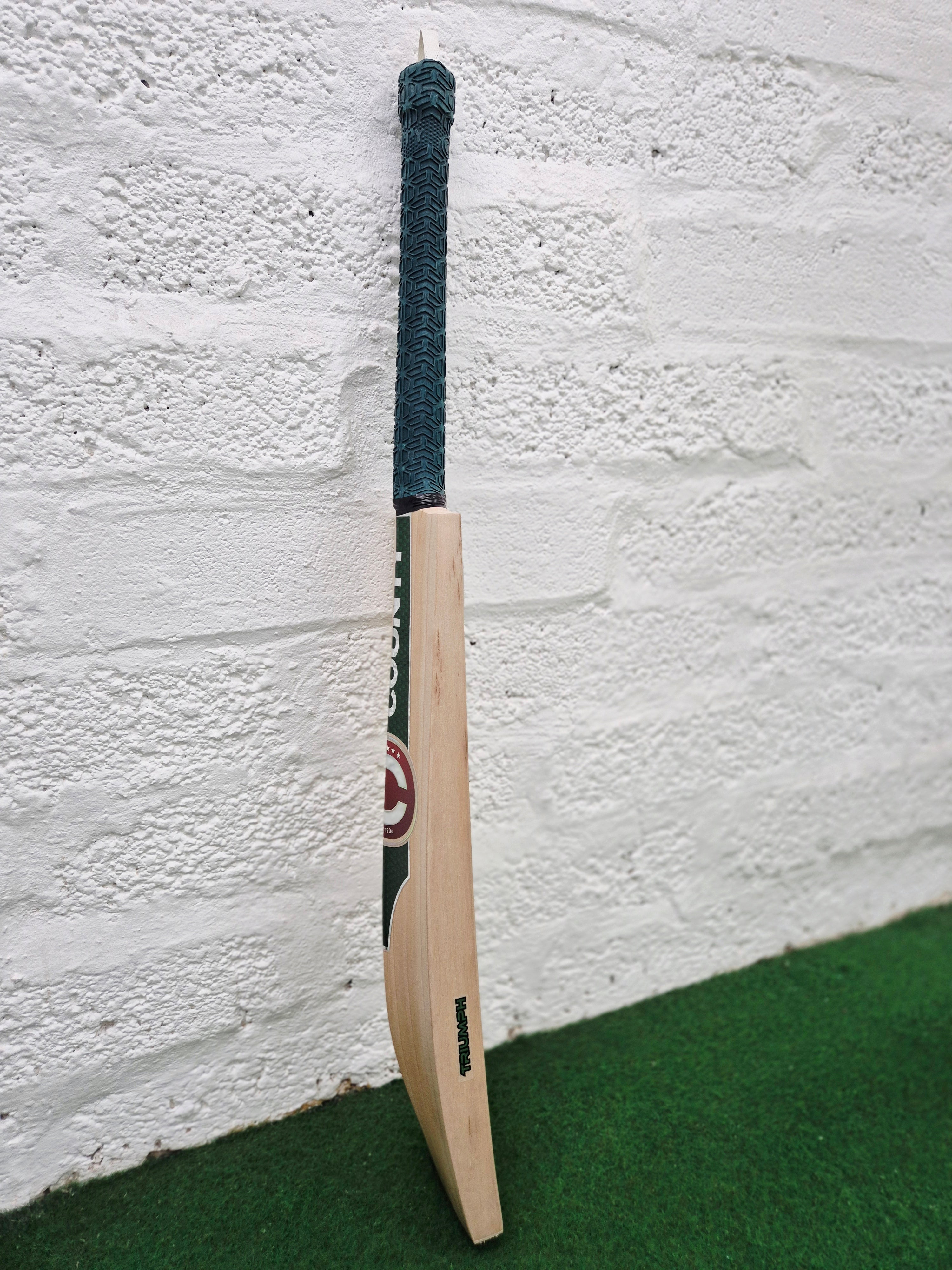 A Hunts County Triumph Senior Cricket Bat by Hunts County, featuring a dark green handle, leans against a white brick wall and rests on a green carpeted surface.