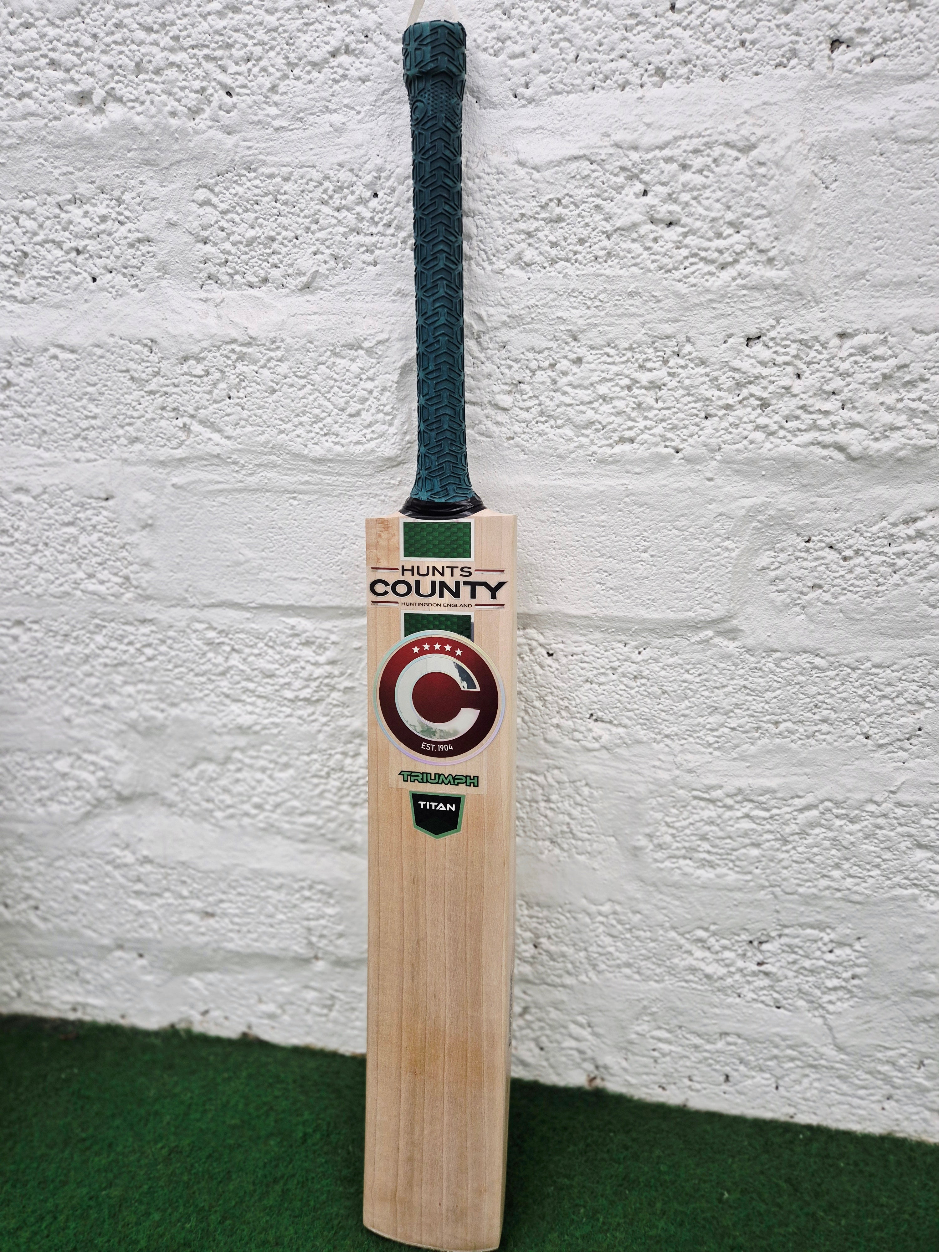 A wooden Hunts County Triumph Senior Cricket Bat with a green handle stands upright against a white textured wall, resting on a green carpet. The bat’s label displays the Hunts County brand, making it an ideal choice for players seeking the quality of a senior cricket bat.