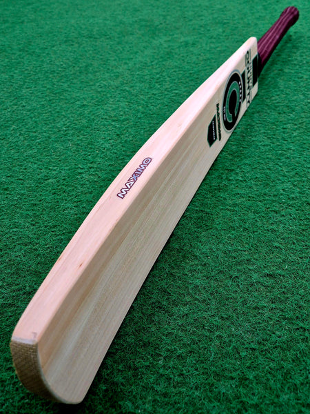 A close-up of the Hunts County Maximo Senior Cricket Bat by Hunts County, featuring a dark handle and resting on a green textured surface.
