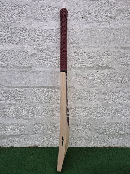 A Hunts County Maximo Senior Cricket Bat by Hunts County, featuring a dark brown grip, is standing upright against a white textured brick wall on a green carpeted surface.