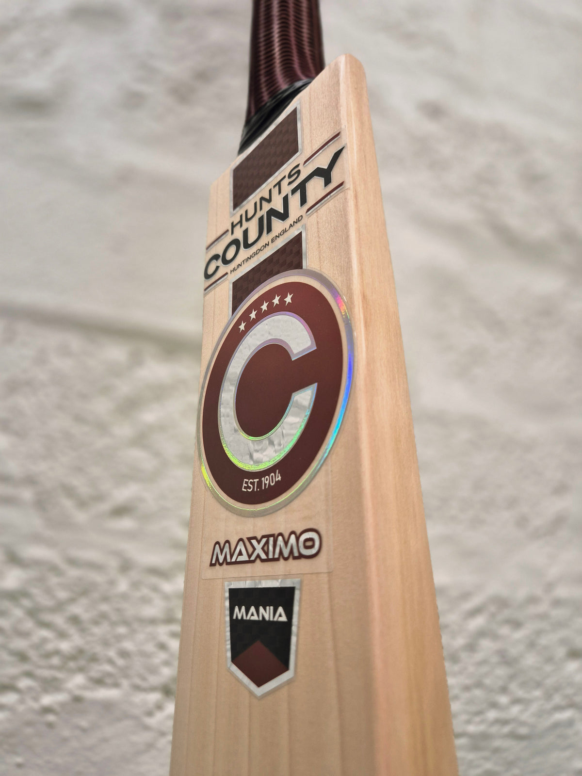 Close-up of the Hunts County Maximo Senior Cricket Bat, crafted from English Willow and showcasing the distinctive Hunts County branding, Mania labels, and a bold C emblem, set against a textured light-colored wall.