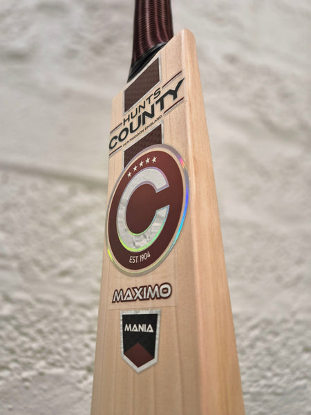 Close-up of the Hunts County Maximo Senior Cricket Bat, crafted from English Willow and showcasing the distinctive Hunts County branding, Mania labels, and a bold C emblem, set against a textured light-colored wall.