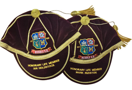 Two maroon velvet Custom Honour & Commemorative Caps by Martin Berrill Sports, each with gold trim, tassels, embroidered crest, and “Hessle C.C.”—one honoring Ian Holdstock and one Mark Rudston as Honorary Life Members.