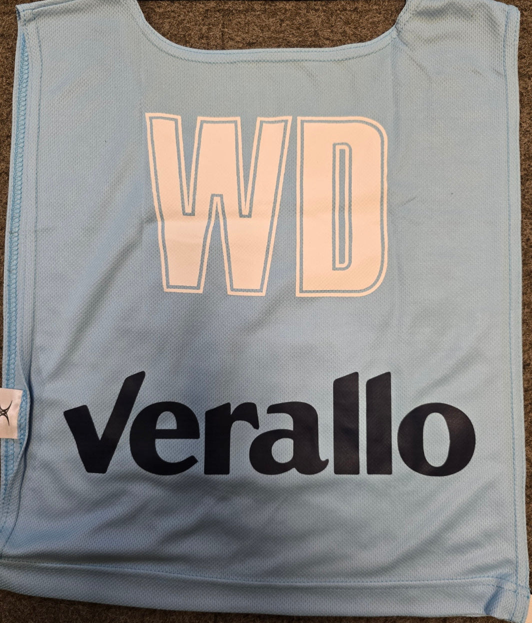 A light blue Personalised Gilbert Netball Bib (Single Bib) by Gilbert shows large white "WD" letters and "Verallo" printed in bold black below, displayed flat on a brown textured surface.