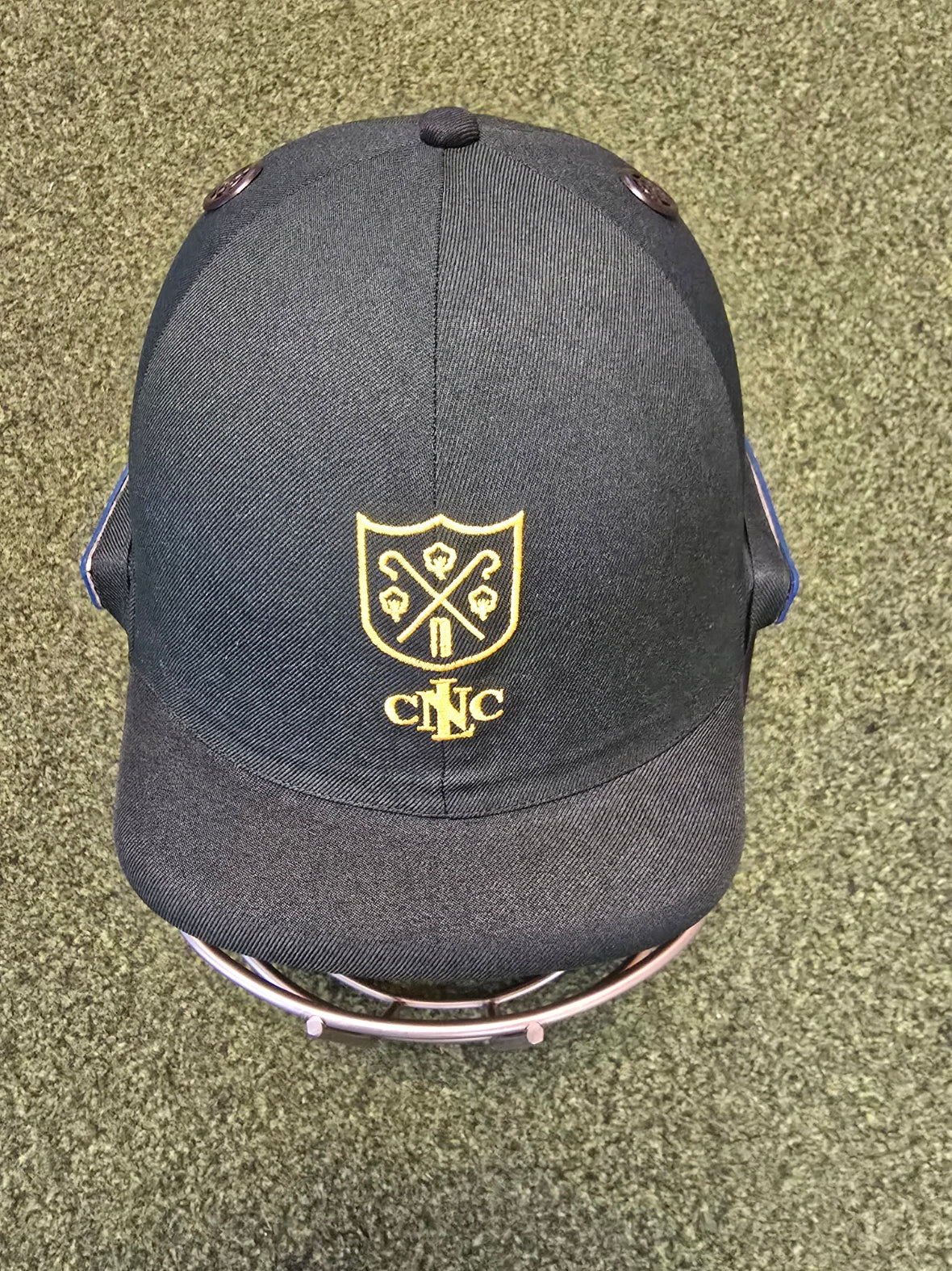A Custom Shrey Cricket Helmet by Shrey, featuring a yellow embroidered club logo with golf clubs and "CNC," reminiscent of designs on black baseball caps, rests on a green textured surface.