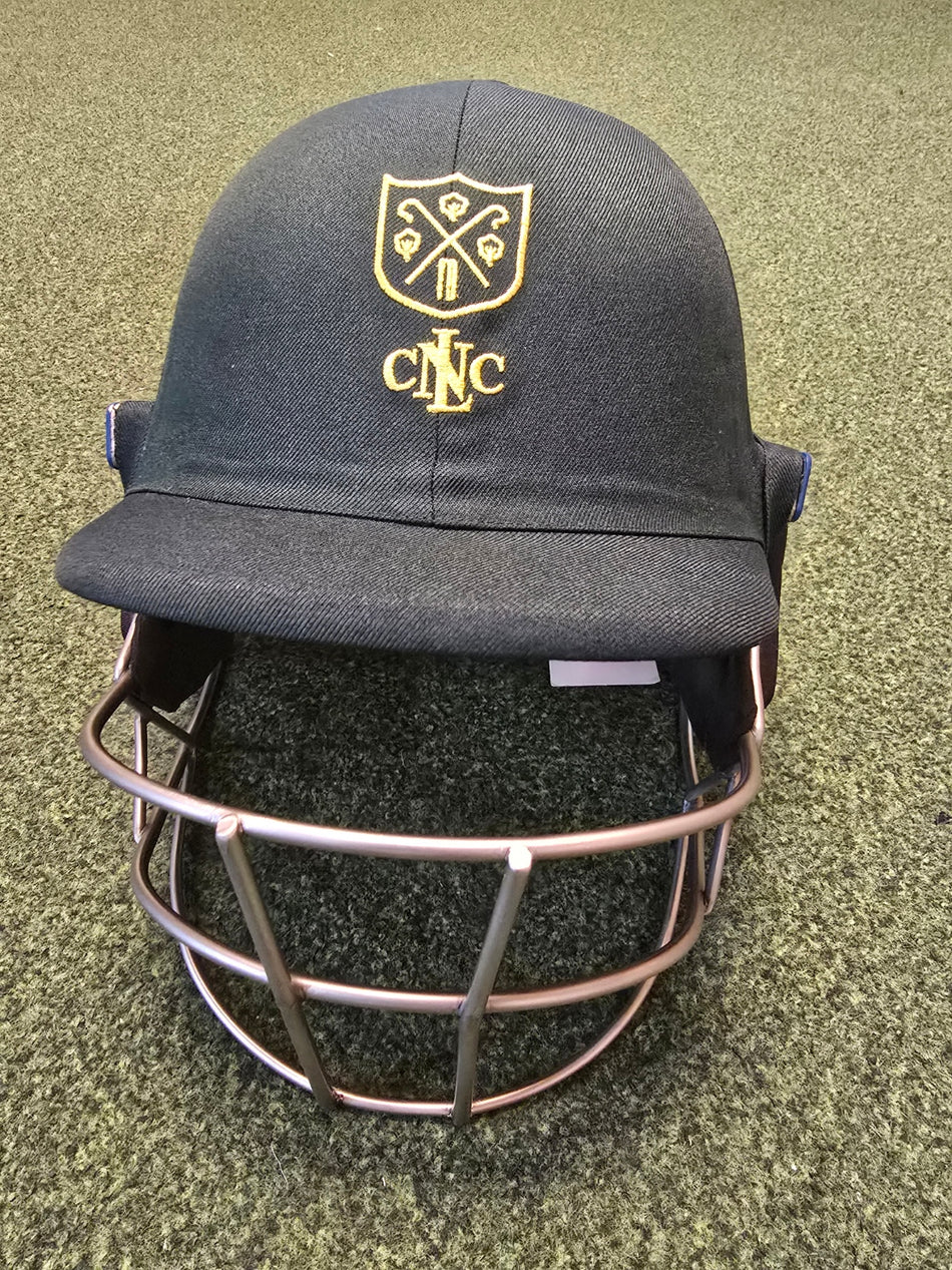 A Custom Shrey Cricket Helmet with a metallic grill, gold embroidered club logo, and CNC letters on the front is shown on a textured green surface.