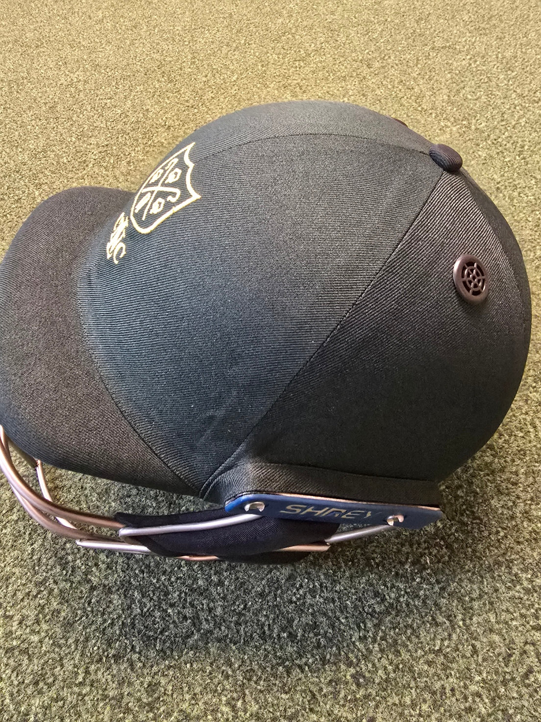 A black baseball cap with a custom embroidered club logo and sunglasses on the brim sits on a speckled green carpet, reflecting the attention to detail found in Custom Shrey Cricket Helmets with Club Logo by Shrey.