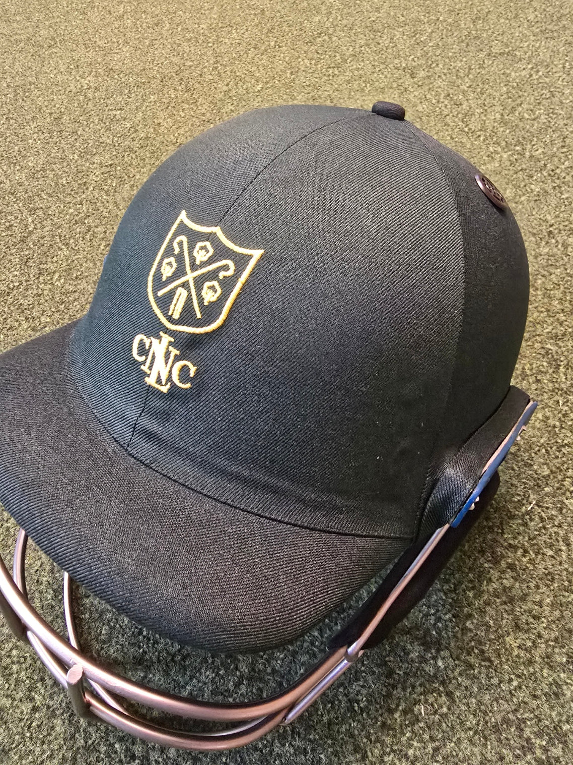 A close-up of a Shrey Custom Cricket Helmet with a gold club logo and "CNC" on the front, resting on a green textured surface.