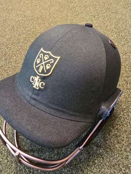 A close-up of a Shrey Custom Cricket Helmet with a gold club logo and "CNC" on the front, resting on a green textured surface.