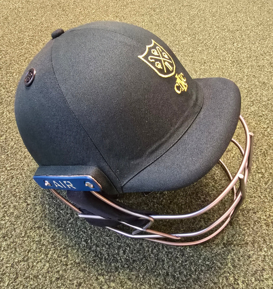 A Custom Shrey Cricket Helmet featuring a gold club logo and CWC embroidery on the front sits on a textured green surface. The navy blue helmet is made by Shrey and includes a durable metal face guard.